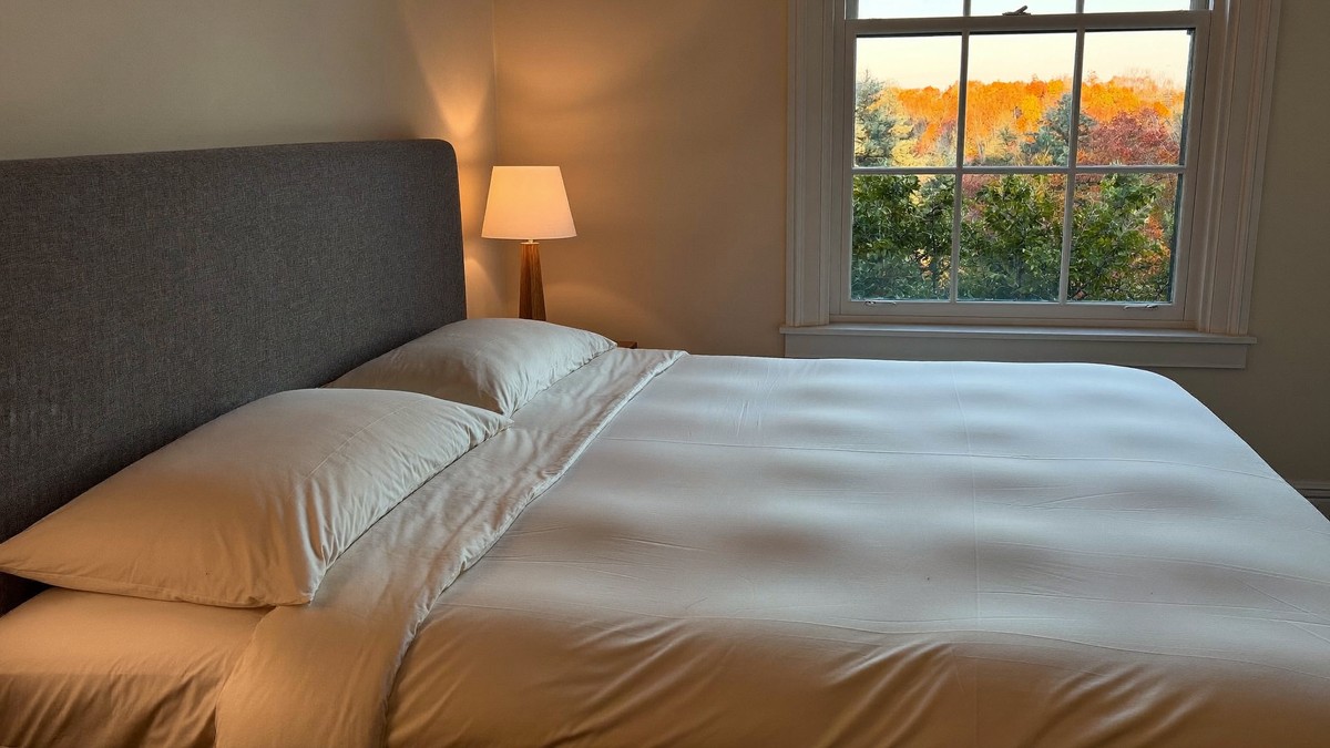 A neatly made double bed with a smooth cream duvet and a grey upholstered headboard in a lamplit bedroom at dusk, with autumn trees through a sash window